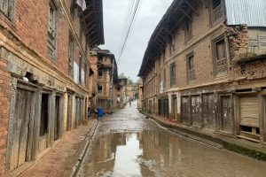 A narrow street lined with old brick buildings features wooden doors and carved window frames, with puddles reflecting the architecture after rainfall. Electrical wires stretch overhead as a few people walk in the distance. 