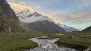 A serene mountain landscape at sunset, with snow-capped peaks and clouds above. A gentle stream meanders through a lush green valley, evoking tranquility.