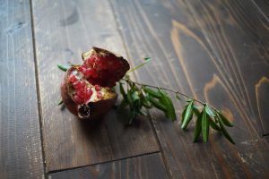 A cracked-open pomegranate with bright red seeds exposed, placed on a dark wooden surface next to a small green leafy branch.
