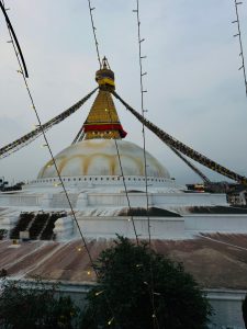 A grand stupa with a golden spire and colorful prayer flags set against a twilight sky. String lights hang in the foreground, adding a serene ambiance.