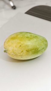 Close-up of a ripe green-yellow mango resting on a white surface, showcasing its smooth skin, natural color variations, oval shape, and fresh, ready-to-eat appearance.
