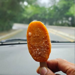A hand holds an orange ice cream bar with frost, set against a blurred car dashboard and a tree-lined road