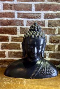 A dark matte bronze Buddha head bust with spiral ushnisha curls and serene downcast expression, displayed on a wooden shelf against an exposed brick wall.