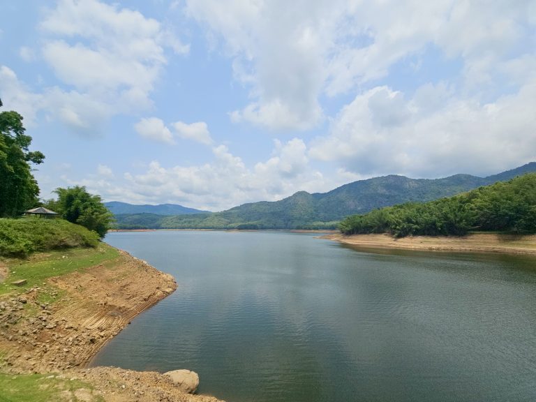 A wide reservoir stretches between green hills under a sky filled with soft clouds. The peaceful scenery showcases the natural beauty of the Western Ghats. Captured at Parambikulam Tiger Reserve, Palakkad.