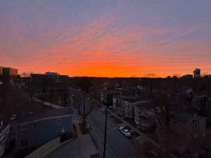 A high-angle, wide landscape shot looking down an empty urban street flanked by houses and bare winter trees. The entire upper half of the frame features a vibrant sunset, with a brilliant band of bright orange fire on the horizon transitioning into a wide pattern of pink and purple rippled clouds. 