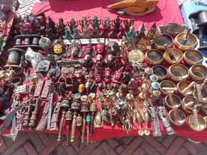 A vibrant market stall overflowing with traditional Buddhist and Hindu crafts including singing bowls, deity figurines, prayer wheels and brass sculptures on a red cloth