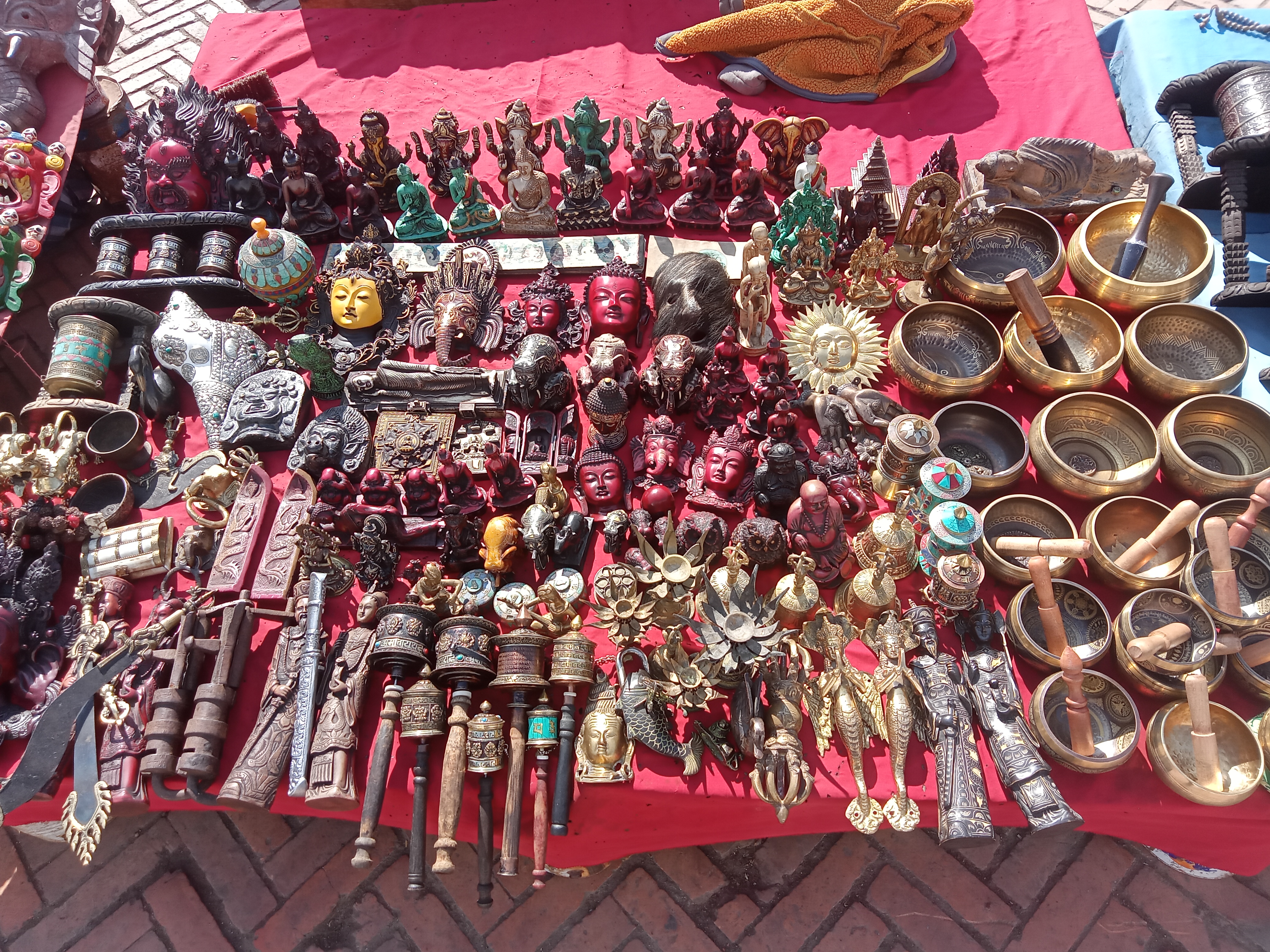A vibrant market stall overflowing with traditional Buddhist and Hindu crafts including singing bowls, deity figurines, prayer wheels and brass sculptures on a red cloth