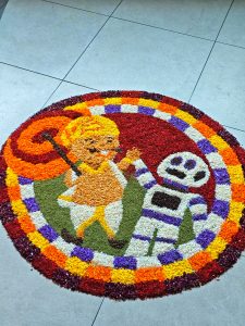 Colorful floral rangoli on a tiled floor depicts a child in a yellow outfit with a walking stick and a robot. Vibrant reds, oranges
