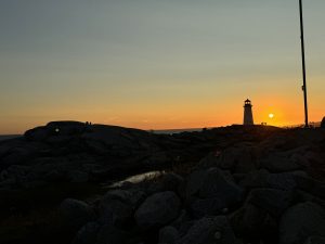 Wide landscape of a lighthouse silhouetted against a brilliant orange sunset. A round white sun sits low on the horizon. The foreground features massive dark coastal boulders in deep shadow, where tiny silhouettes of people stand near the lighthouse and on a ridge. A thin dark pole frames the far right. 