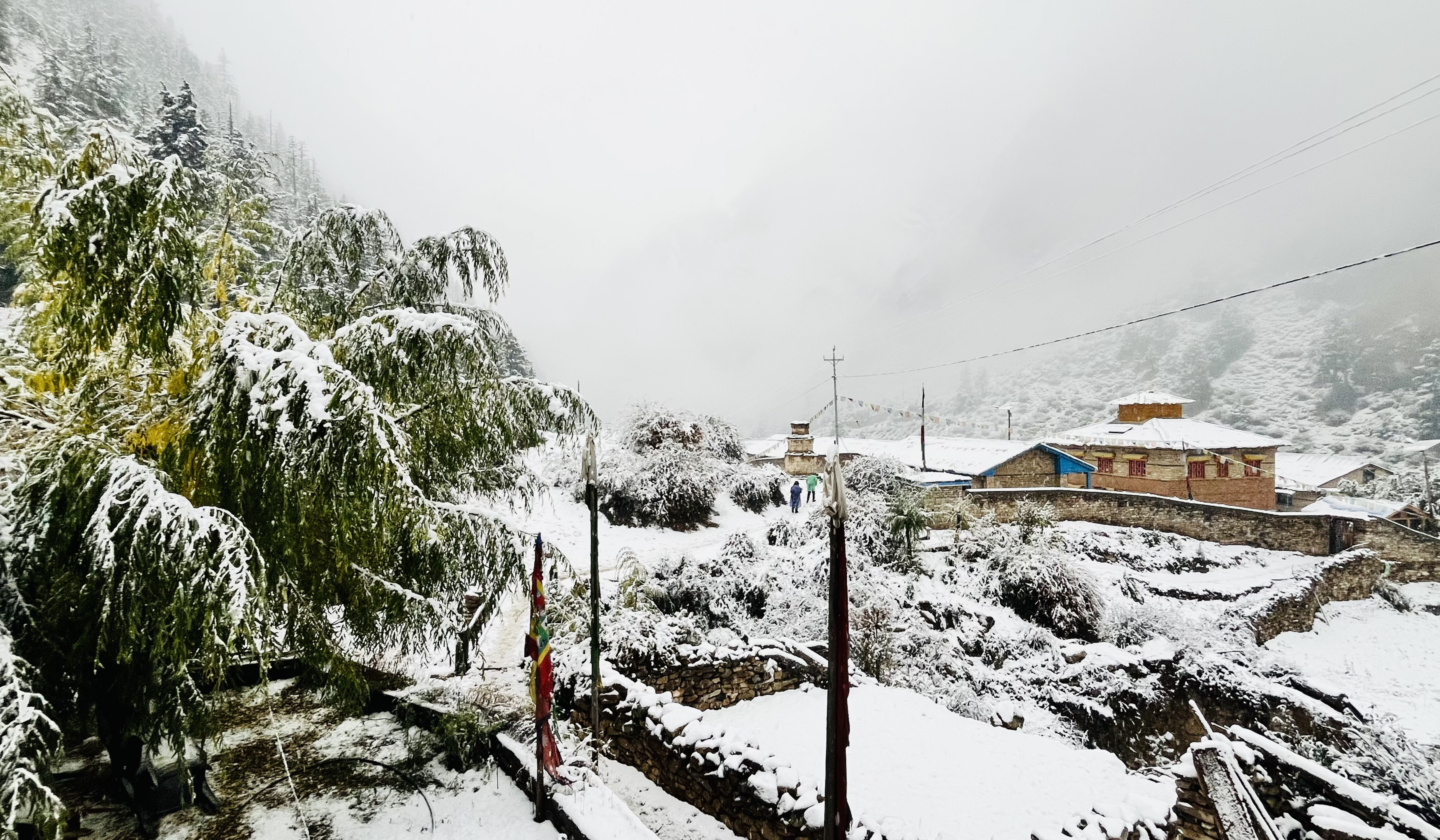 A mountain village with stone houses and snow‑covered roofs sits among terraced hills and trees blanketed in fresh snow, with mist obscuring the higher peaks. A few people walk along a snowy path.