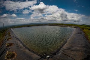 A large, rectangular water reservoir is depicted, surrounded by a black, plastic lining at its edges. The water is calm and reflects the sky, which is partly cloudy with patches of blue peeking through.