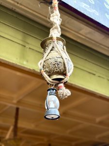 Ornate brass Nepali Kalash with relief carvings, suspended by braided jute rope with colorful thread accents, with a miniature metal lantern hanging below.