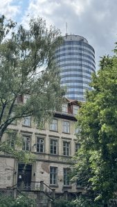 The cylindrical glass Jentower rises behind a weathered historic stone building in Jena, Thuringia. Ornate window frames and faded details contrast with the tower’s reflective facade, as lush trees and a partly cloudy sky frame the scene.
