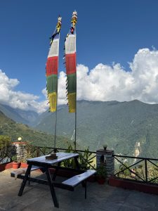 Two tall, colorful prayer flags stand beside a wooden picnic table on a terrace, overlooking a vast, green mountain landscape under a bright blue sky.