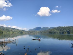 Lone fisherman rowing across a glassy lake with fish traps and green mountain reflections 