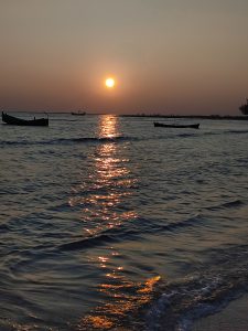 A calm ocean sunset with an orange sun near the horizon, golden light reflecting on gentle waves, with distant boats and a faint shoreline in silhouette.
