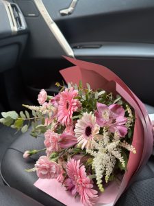A bouquet of flowers, featuring various shades of pink, is placed on a textured black car seat. The bouquet includes large, vibrant pink gerbera daisies, soft pink roses, and delicate white flowers with fine, feathery textures.