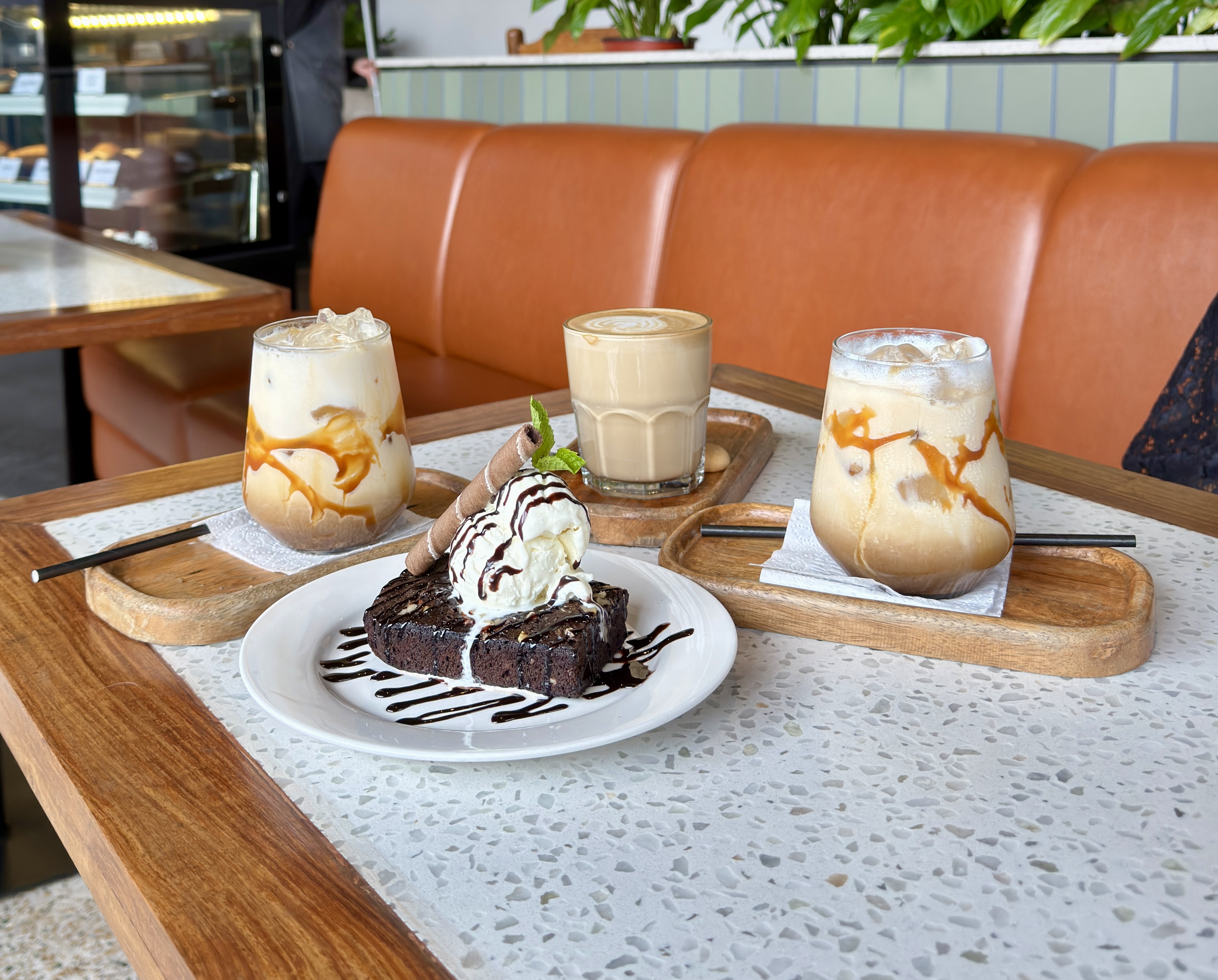 A cafe table with a chocolate brownie topped with vanilla ice cream and chocolate sauce, flanked by two iced caramel lattes and a hot latte with latte art, served on wooden trays at a modern cafe in Nepal.