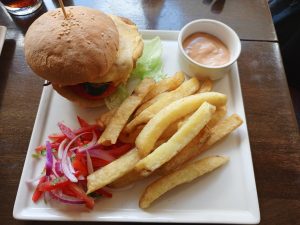 A square white plate holds a cheeseburger with lettuce, tomato, and an olive slice, secured with a wooden toothpick. A large portion of French fries occupies the center, accompanied by a small white bowl of pink dipping sauce and a side salad of chopped red onions, tomatoes, and parsley. 