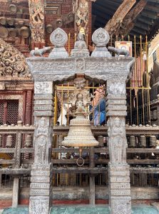 An ornate stone bell tower with a large hanging brass temple bell suspended by chains, flanked by deity figures and topped with Buddhist medallions, at a Newari temple complex, Nepal. 