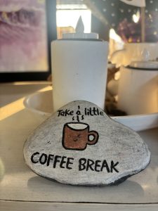 Smooth stone with a painted mug and text saying "Take a little coffee break." It's placed on a table with soft lighting and a white teapot in the background.