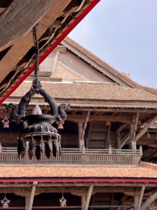 Ornate bronze lamp with intricate dragon design hanging from a wooden beam, under a red-tiled roof with traditional architectural elements.