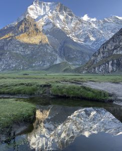 Snow-covered mountain peaks under a clear blue sky are reflected in a calm water body below, surrounded by lush green grass, creating a serene scene.