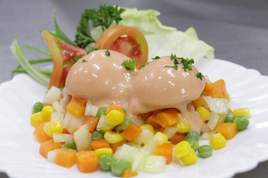 Two rounded scoops of a creamy salad mixture topped with chopped parsley, served on a bed of mixed diced vegetables including carrots, peas, corn and white vegetables, with lettuce and sliced tomato garnish on a white plate.