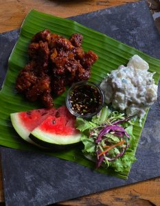 Top-down view of a meal served on a vibrant green banana leaf over a dark slate. It features savory, sauce-coated meat pieces, a creamy potato salad side, a crisp mixed green salad, two small watermelon slices, and a tiny bowl of dipping sauce.
