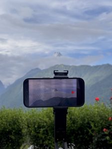 A peaceful environment, where timelapse is being captured in phone where it shows clouds, mountain peak and greenery in background.
