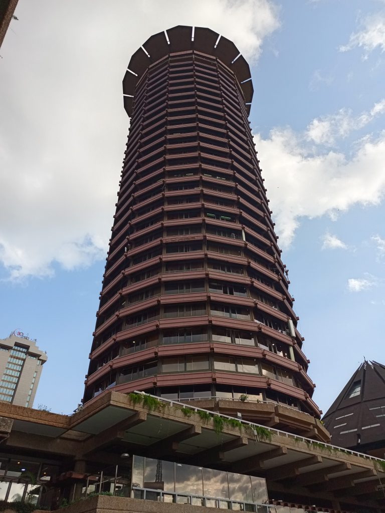 A high-angle view looking up at KICC a tall, circular skyscraper with a brown multi-tiered facade under a partly cloudy sky.