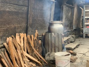 A large metal pot sits on a makeshift stove made of corrugated sheets, with smoke rising from burning wood underneath. Beside the stove, a pile of split firewood is stacked against a rustic wooden wall, and a white plastic bucket rests on the stone ground.
