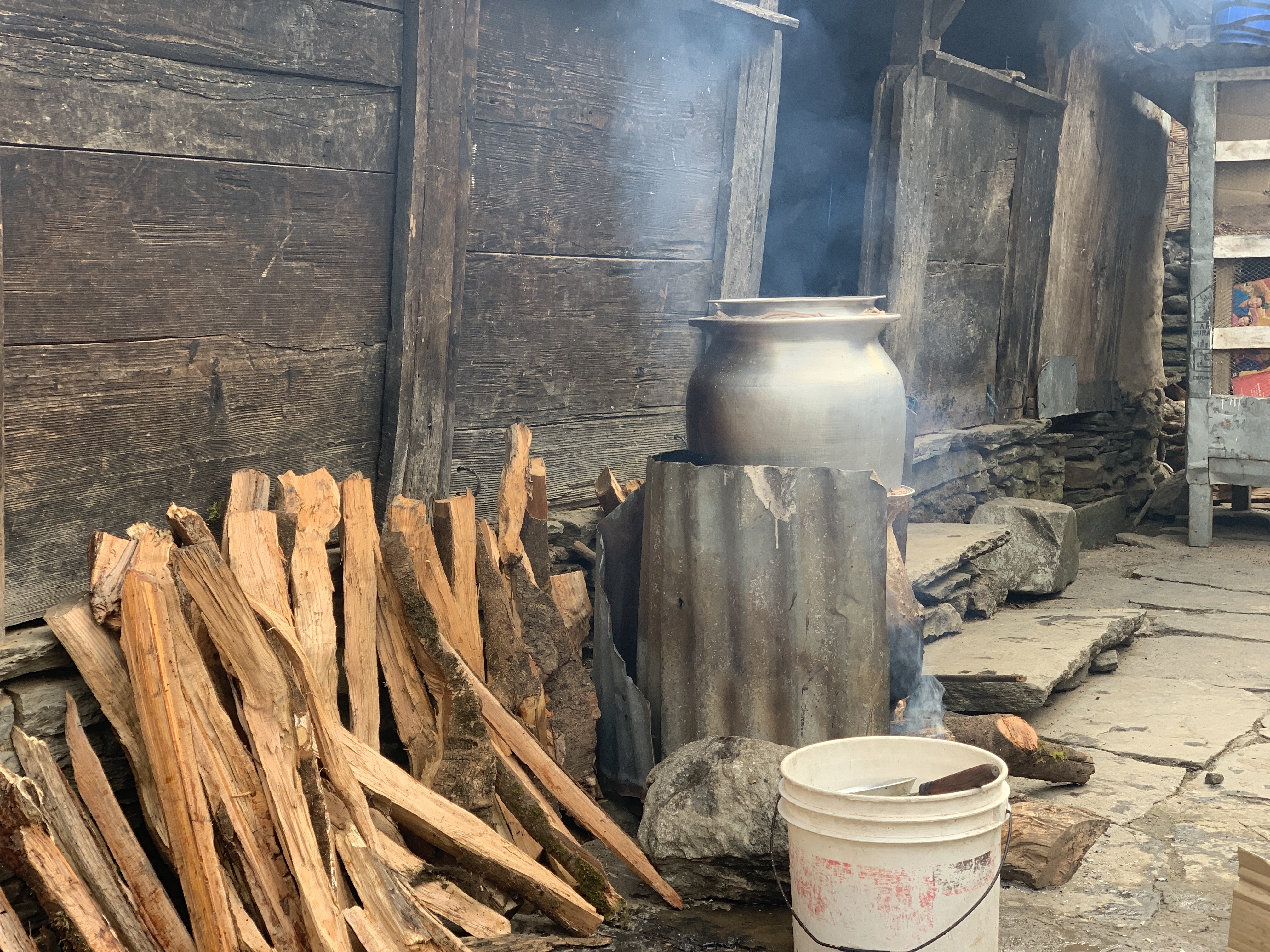 A large metal pot sits on a makeshift stove made of corrugated sheets, with smoke rising from burning wood underneath. Beside the stove, a pile of split firewood is stacked against a rustic wooden wall, and a white plastic bucket rests on the stone ground.