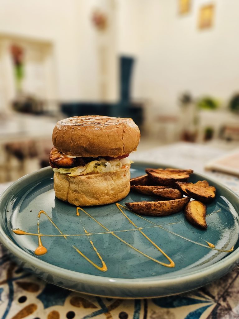 A delicious burger accompanied by golden potato wedges is presented on a blue plate with artistic sauce decoration. The blurred café interior creates a clean background that draws attention to the food. Captured in Kolkata, West Bengal.