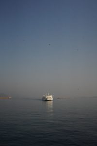 A white ferry sailing on calm blue-gray water under a clear blue sky, with a harbor breakwater and small red lighthouse visible in the distance.

