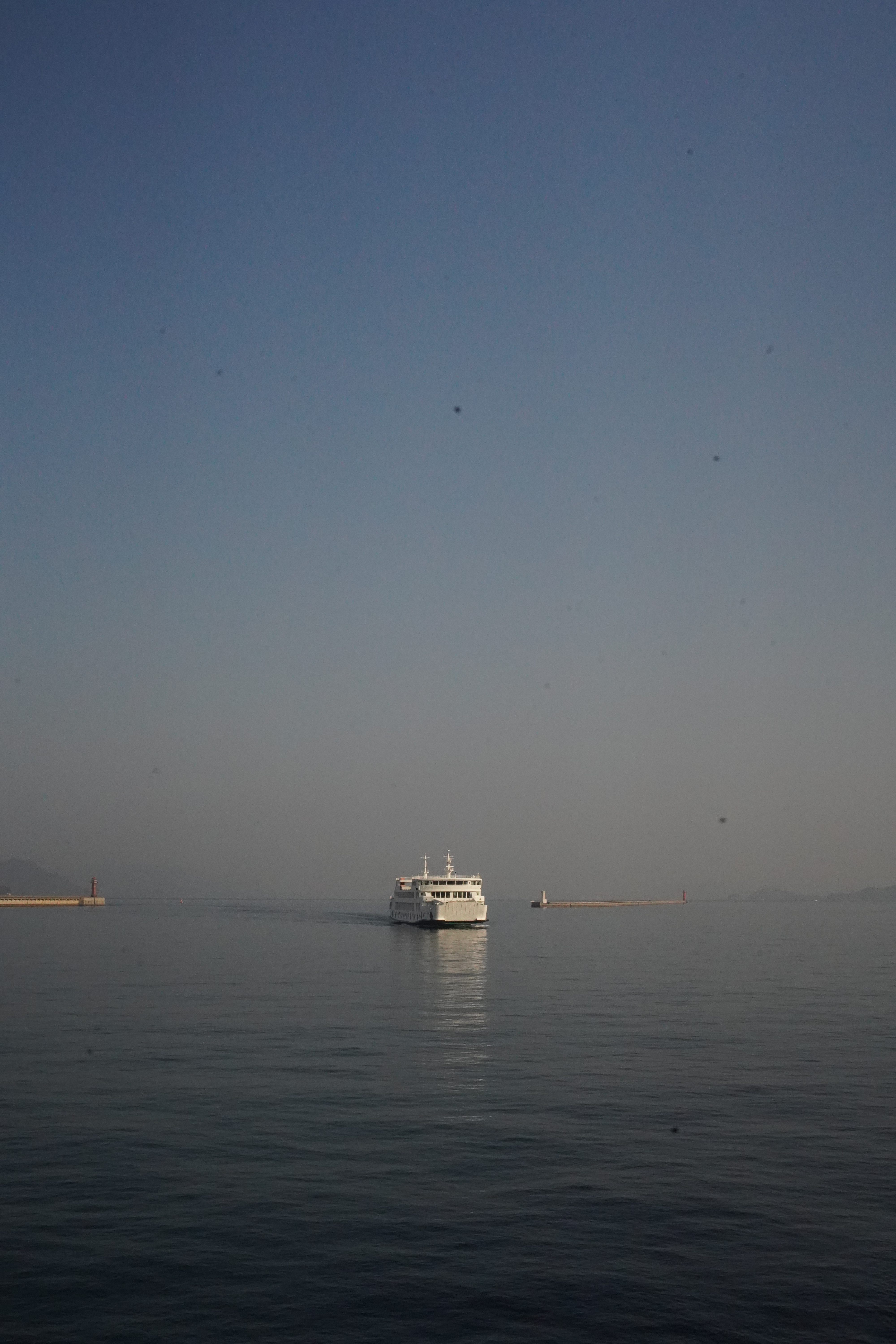 A white ferry sailing on calm blue-gray water under a clear blue sky, with a harbor breakwater and small red lighthouse visible in the distance.