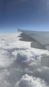 A view from an airplane window showing the large silver wing of an aircraft stretching across the right side of the frame. Below, a massive expanse of dense, billowy white clouds blankets the earth under a bright blue sky.