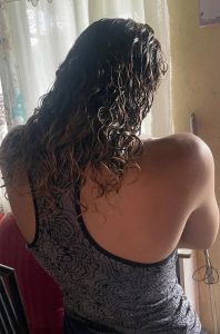 A medium shot showing a person from behind with long, very curly dark brown hair featuring caramel-toned highlights at the ends. They are wearing a gray camisole with a textured black graphic pattern.

