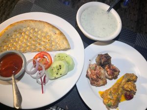 A white plate with naan, sliced onions, cucumbers, tomatoes, and a dipping sauce. Another plate holds grilled meats.