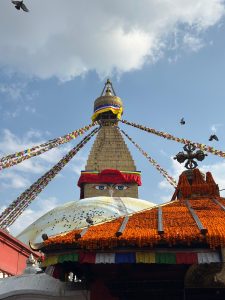 #NepalPhotoFestival2026
Stupa with colorful prayer flags reaching from the golden spire. Buddha eyes and pigeons atop. Bright orange marigolds adorn lower structures. Sky is partly cloudy.