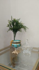 A glass vase with lush green foliage sits on a glass table next to an ornate blue and gold decorative container and a transparent glass jar.