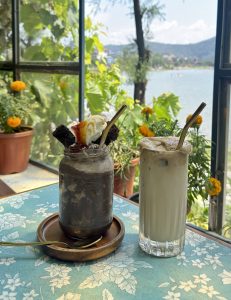 Two specialty cold drinks — a dark chocolate brownie jar shake and a creamy iced coffee — served with metal straws on a floral tablecloth with a view of Phewa Lake and mountains through the window, Pokhara, Nepal. 