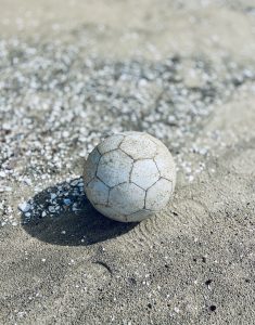 A weathered soccer ball lies on a sandy beach, speckled with seashell fragments