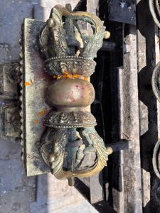 Close-up of an ornate brass vajra (thunderbolt/dorje) ritual object decorated with marigold petals and red sindoor powder, mounted on a temple gate, Nepal.