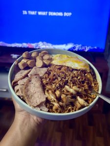 A hand holding a bowl of spicy dry instant noodles topped with a fried egg, sliced processed meat, sausages, and chicken pieces, with a TV screen showing subtitles in the background. 
