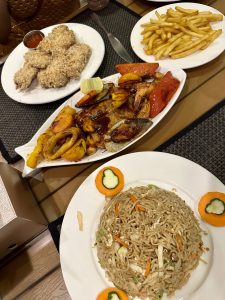 A table with diverse dishes: crispy breaded chicken with dip, golden fries, grilled seafood platter, and noodles with carrot slices, creating a lively feast.