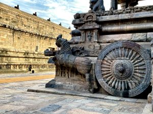 Ancient stone carving of a chariot with an ornately detailed wheel and horse motif, set against a historic temple wall under a clear blue sky.
