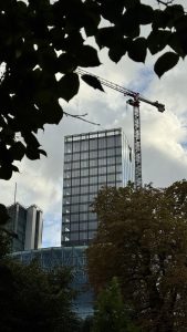 A tall glass skyscraper reflects a dramatic cloudy sky, with a large construction crane rising to its right. Green foliage with hints of autumn color frames the foreground, while a second building peers through the trees behind.
