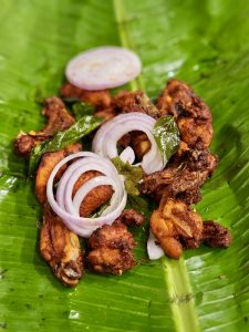 A serving of Kerala-style fried chicken is presented on a banana leaf, garnished with onion rings and fresh curry leaves. The crispy coating, vibrant colors, and traditional presentation create an appetizing food scene that highlights local flavors. Captured in Perumanna, Kozhikode..