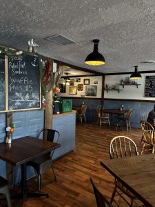 Interior of a cozy tavern with wooden floors and tables. A large chalkboard on the left lists specials, including a pulled pork sandwich and waffle fries for five dollars. The walls are decorated with a nautical net and framed artwork beneath glowing black dome pendant lights. 